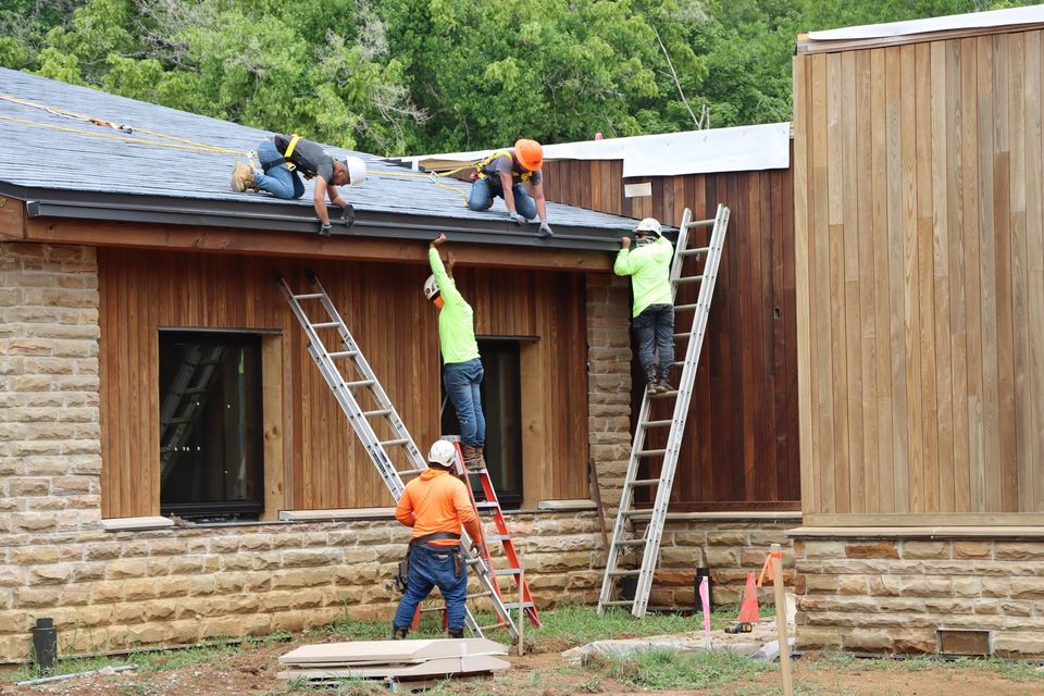 Workers on ladders and the roof edge working on a wood-sided building with stone base